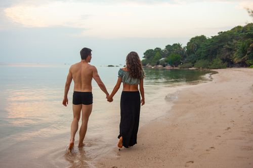 Free A romantic couple walking hand in hand on a serene beach during sunset. Stock Photo