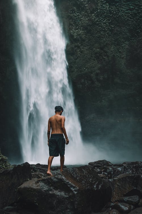 Topless Man in Black Shorts Walking on Rock Near Waterfalls