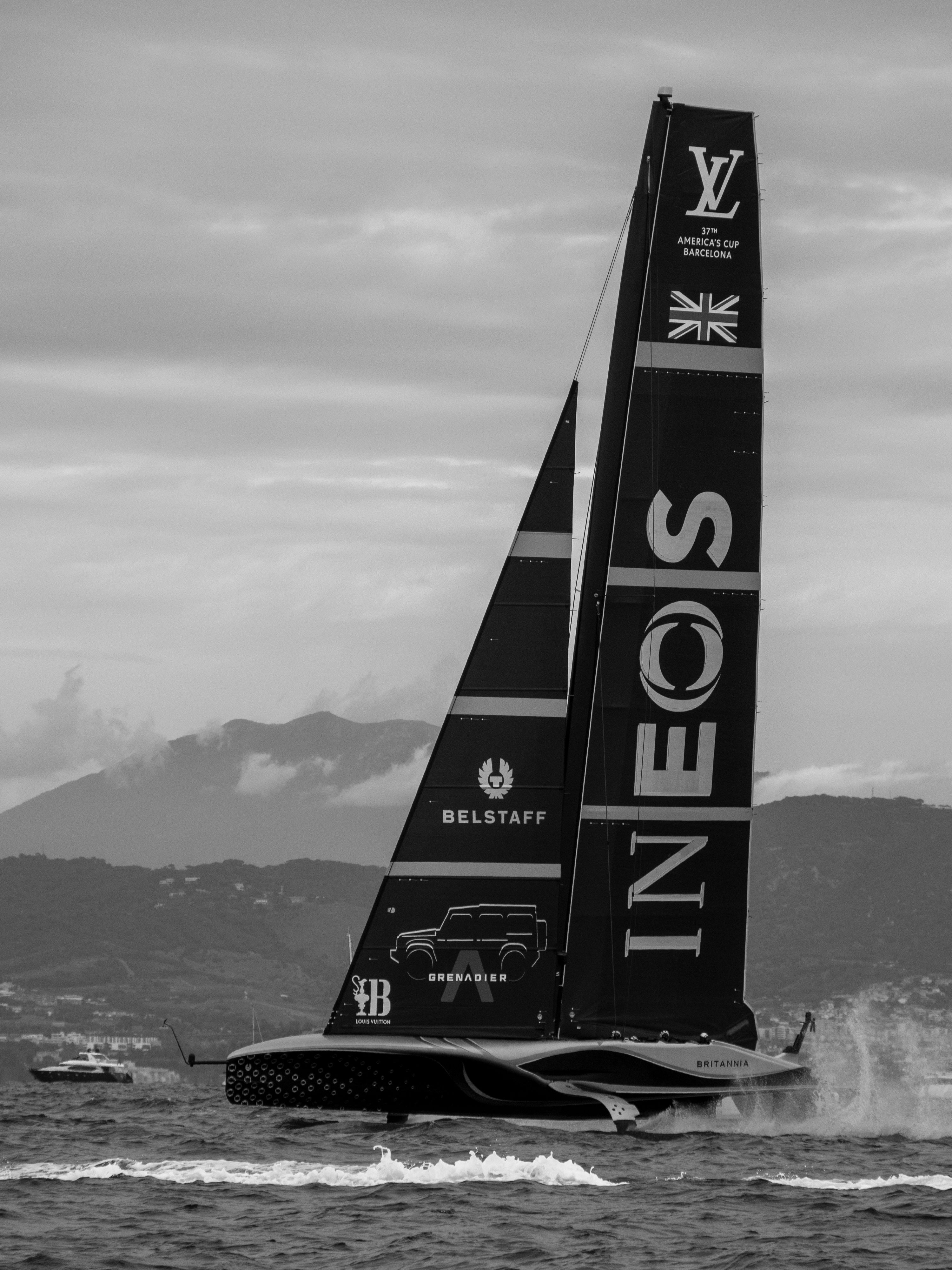 Black and white image of a sleek sailing yacht brandishing INEOS and UK flag, gliding across water.