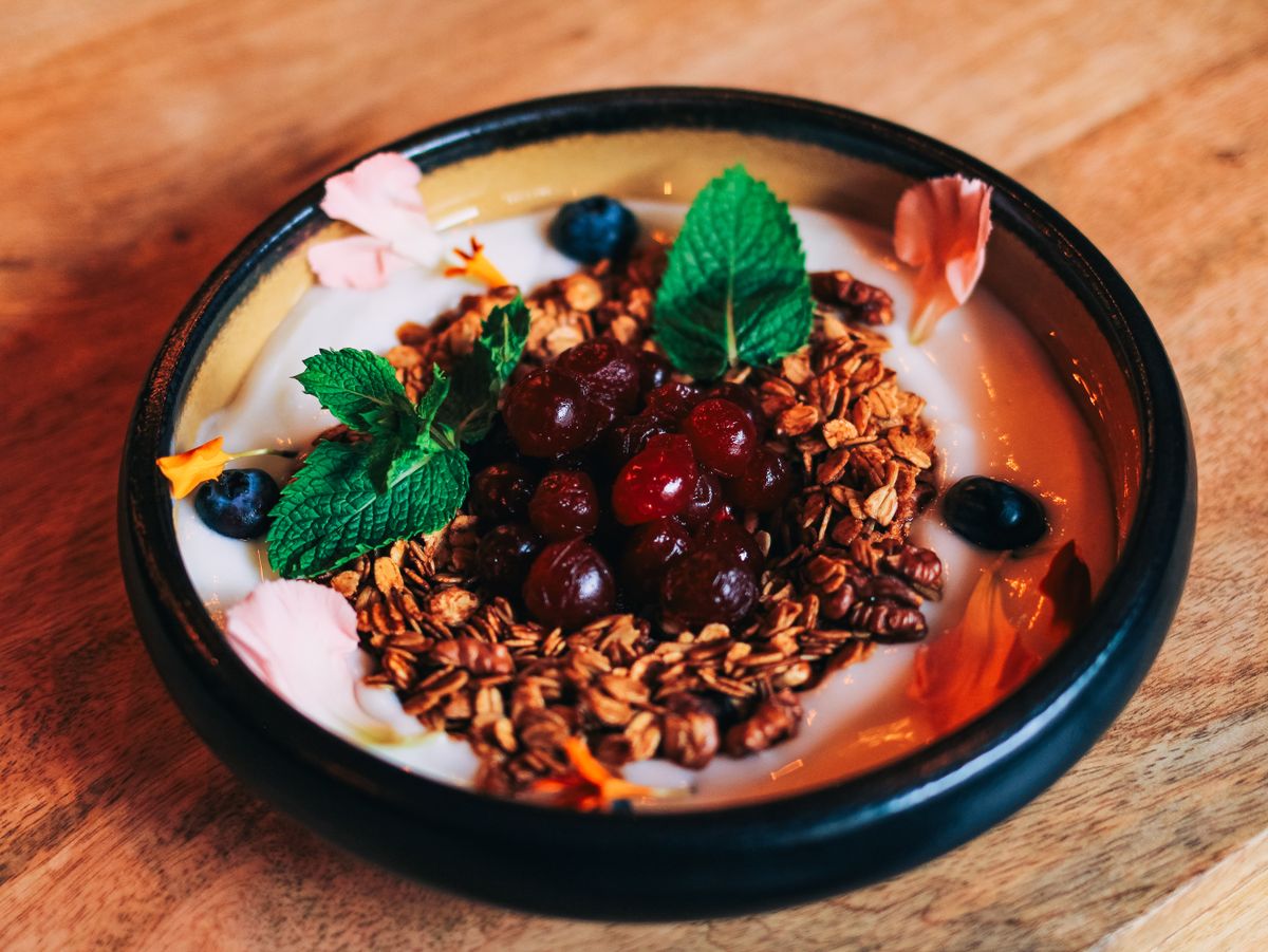 Thick yogurt topped with granola, dark cherries, blueberries, mint leaves and flower petals