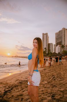 A young woman poses on the beach in Puerto Vallarta during a vibrant sunset, with city skyline in the background.