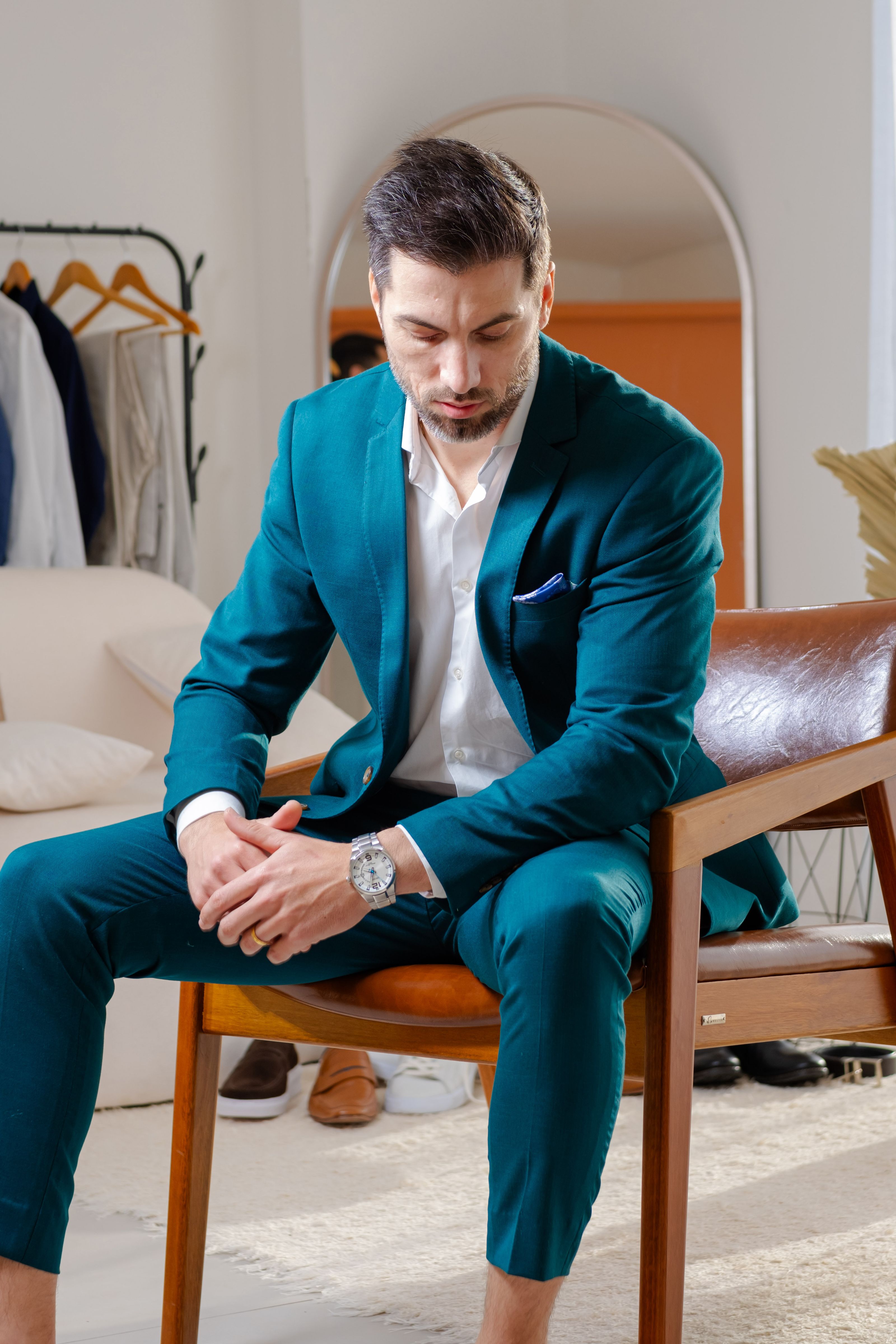 Homem de terno azul-petróleo sentado em um interior moderno, exalando uma presença elegante e ponderada.