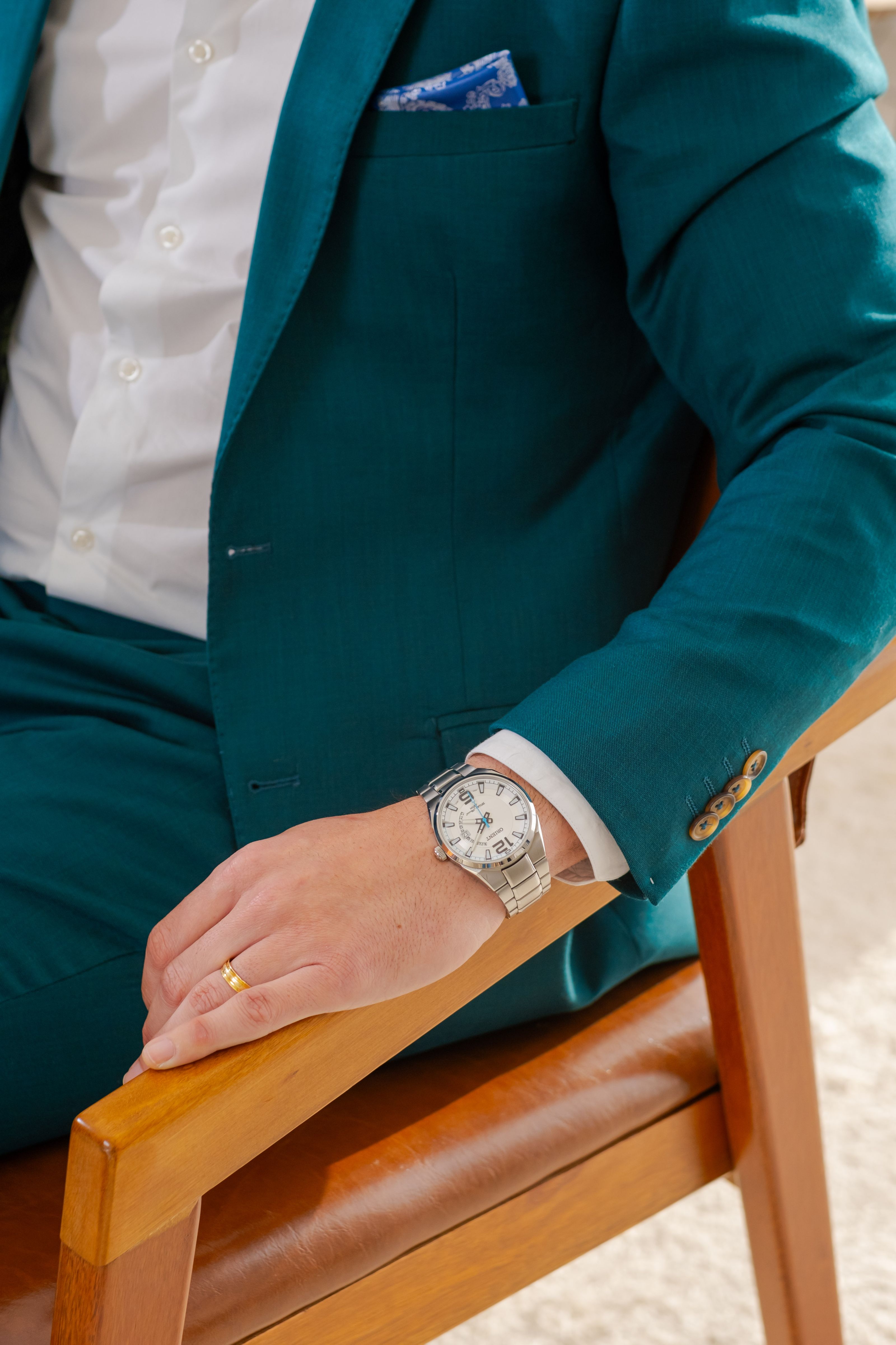 Close-up de um homem elegante vestindo um terno azul-petróleo e um relógio de luxo prateado, sentado em uma cadeira de madeira.