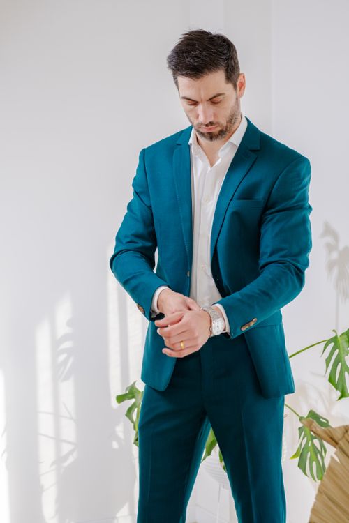 Free A stylish man in a teal suit adjusts his watch with greenery in the background. Stock Photo