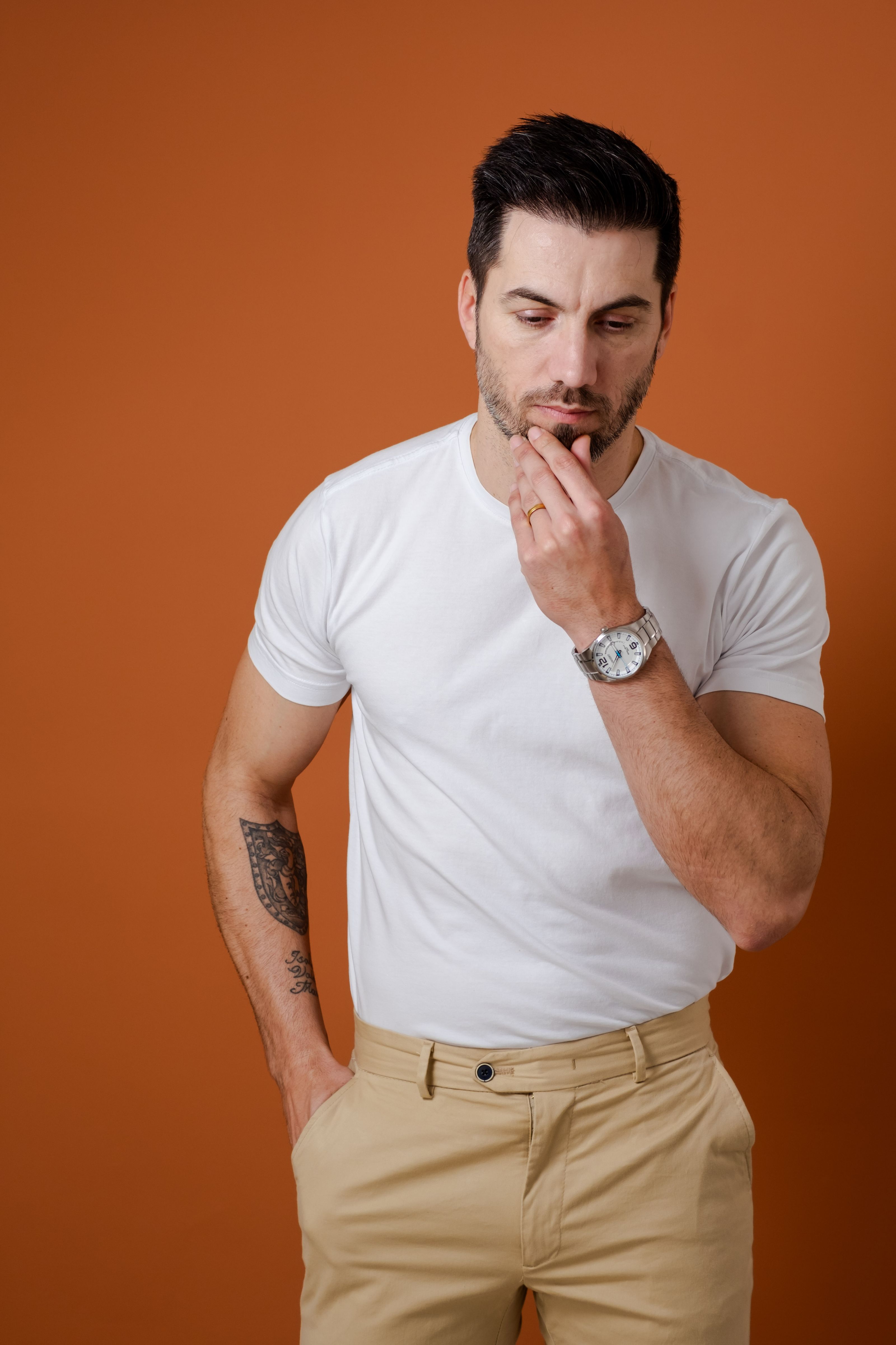 Homem de camisa branca e calça bege pensativo, tendo como pano de fundo laranja.