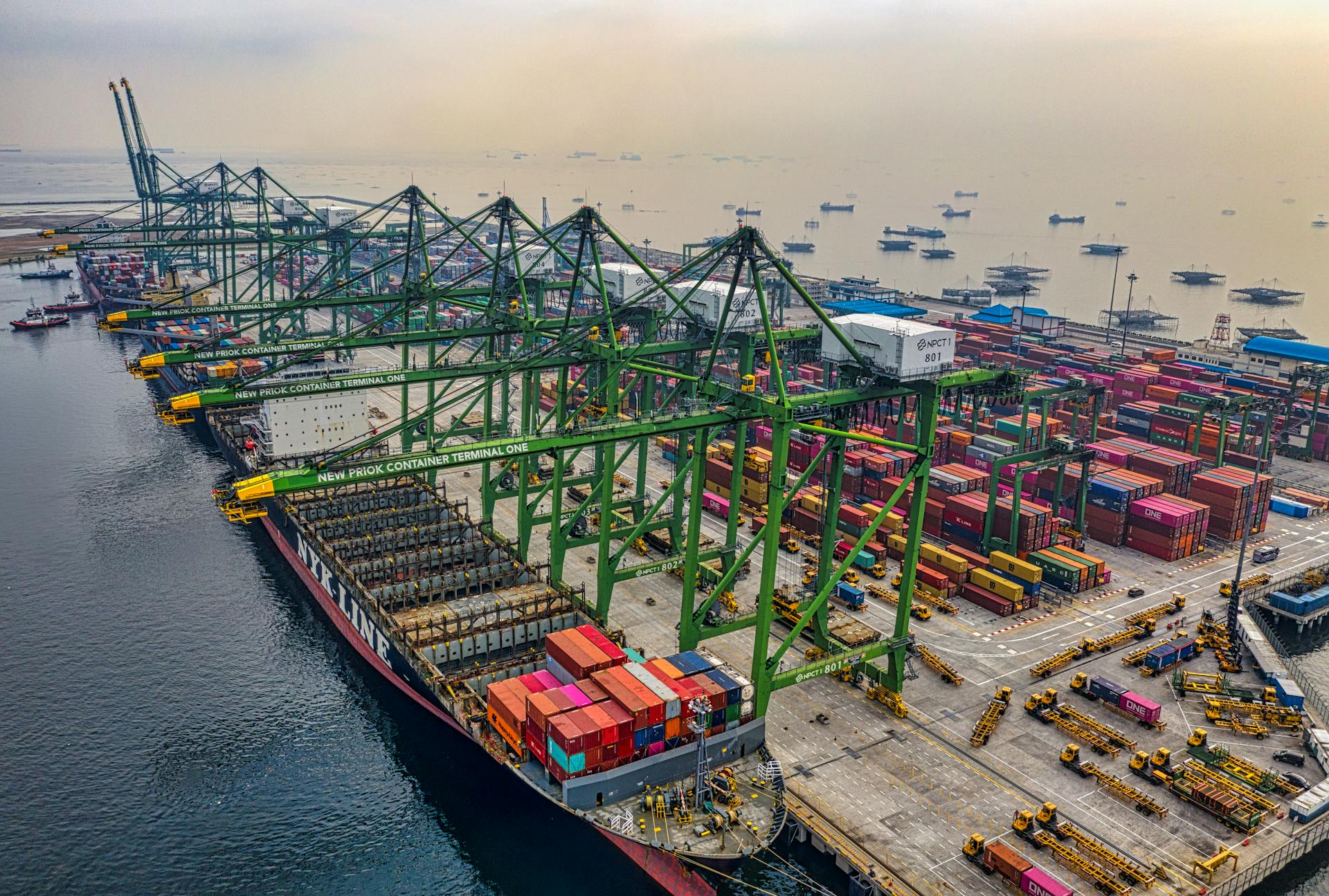 Busy cargo port in North Jakarta with cranes and container ships under a hazy sky.