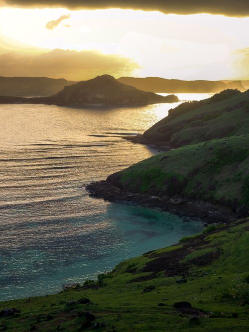 Free Stunning view of the sun setting over a coastal landscape with lush green hills and calm waters. Stock Photo