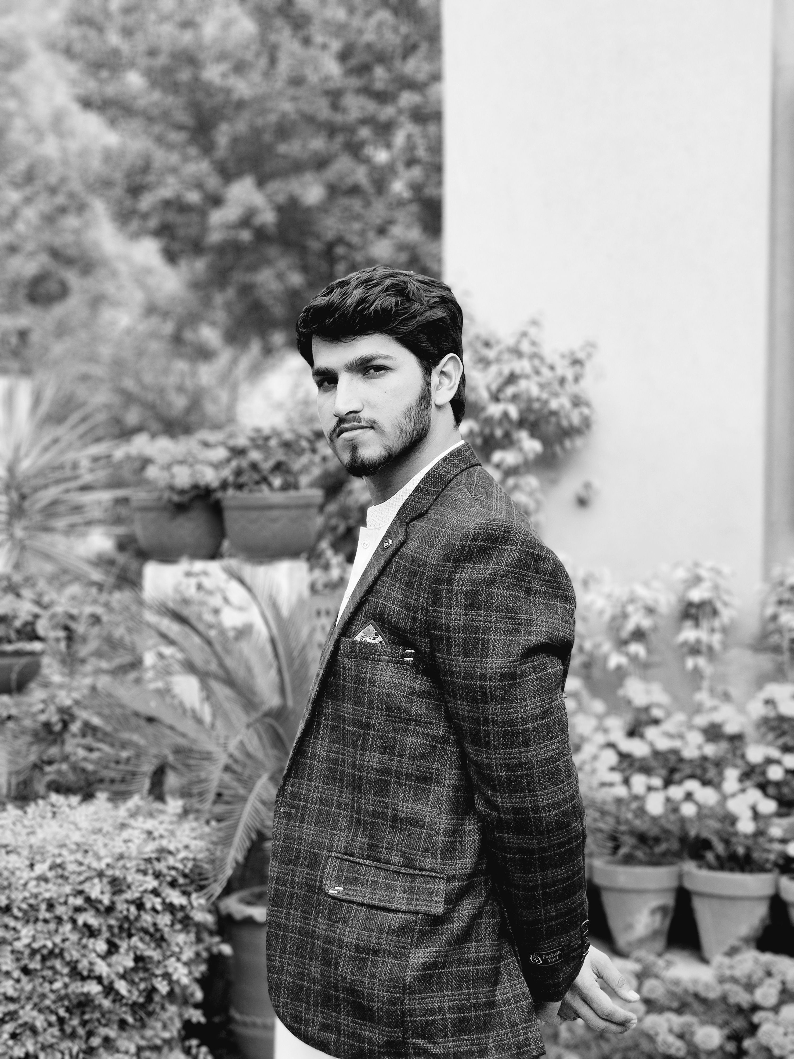 A man in a checkered suit poses in a garden setting. Black and white photo enhancing contrast.