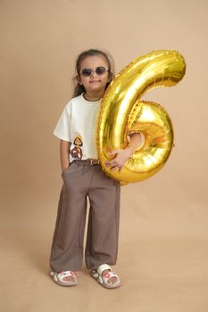 A stylish child holding a gold number six balloon with a neutral background, wearing sunglasses and casual attire.