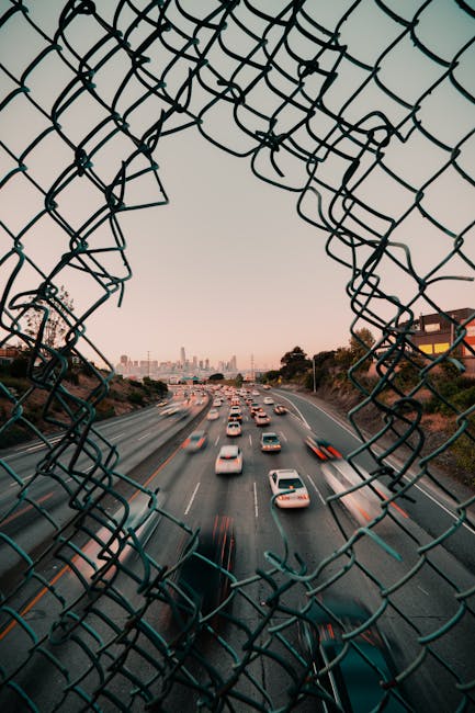 Traffic flows on a highway framed by a broken fence with San Francisco cityscape at sunset.