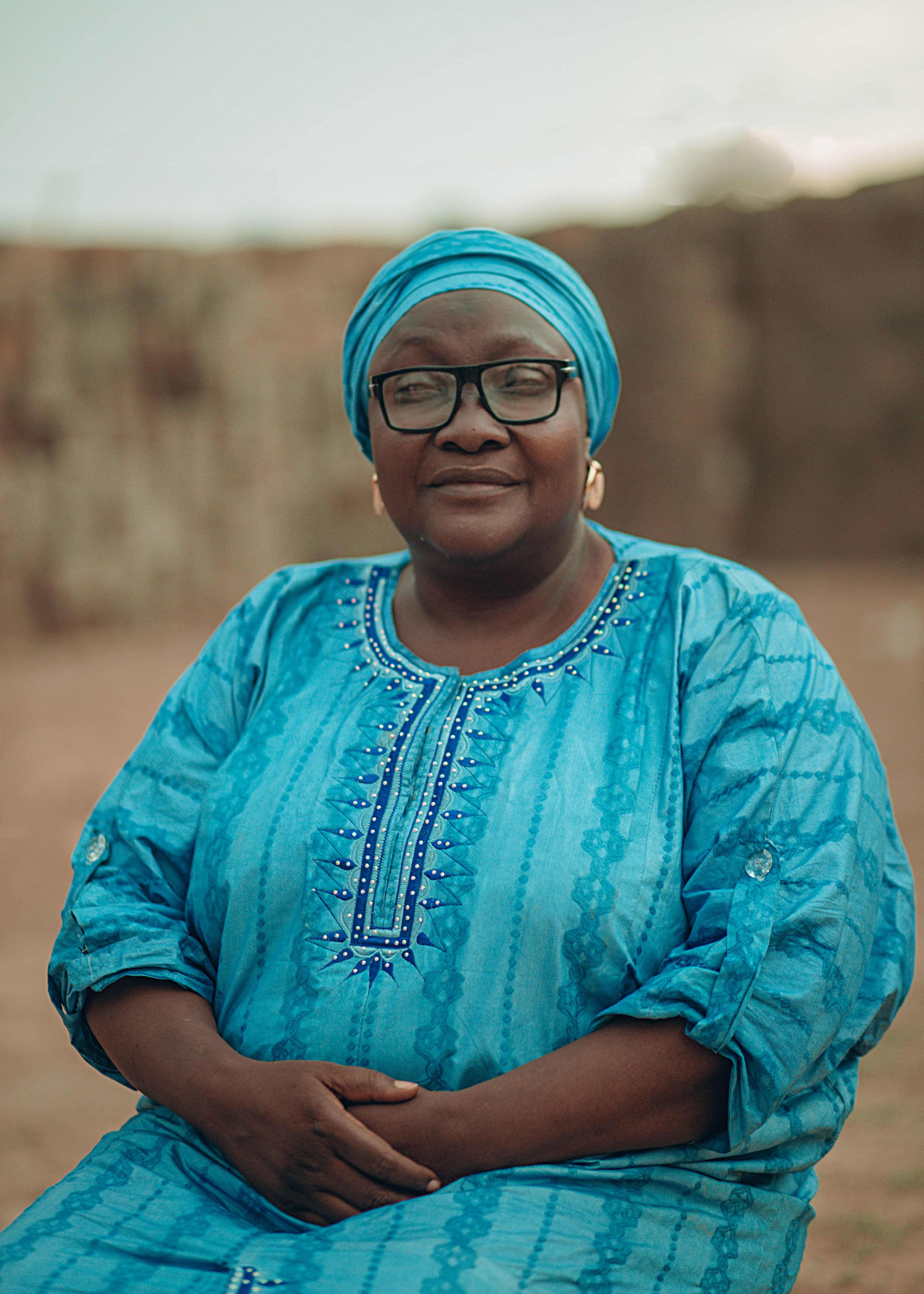An elderly African woman wearing traditional blue attire and glasses, posed outdoors.
