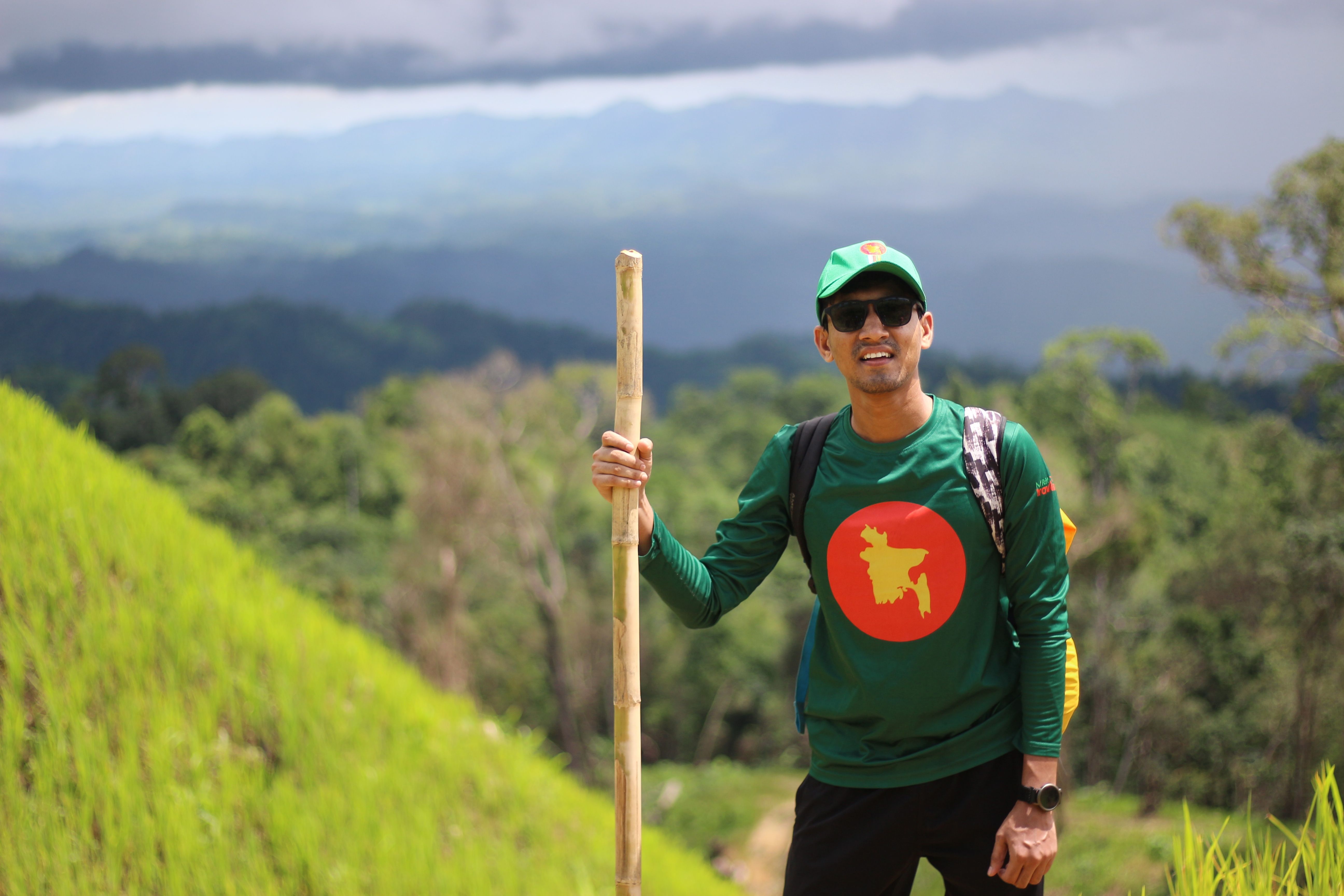 A man wearing a cap and sunglasses hikes in a scenic lush green area with a bamboo stick.