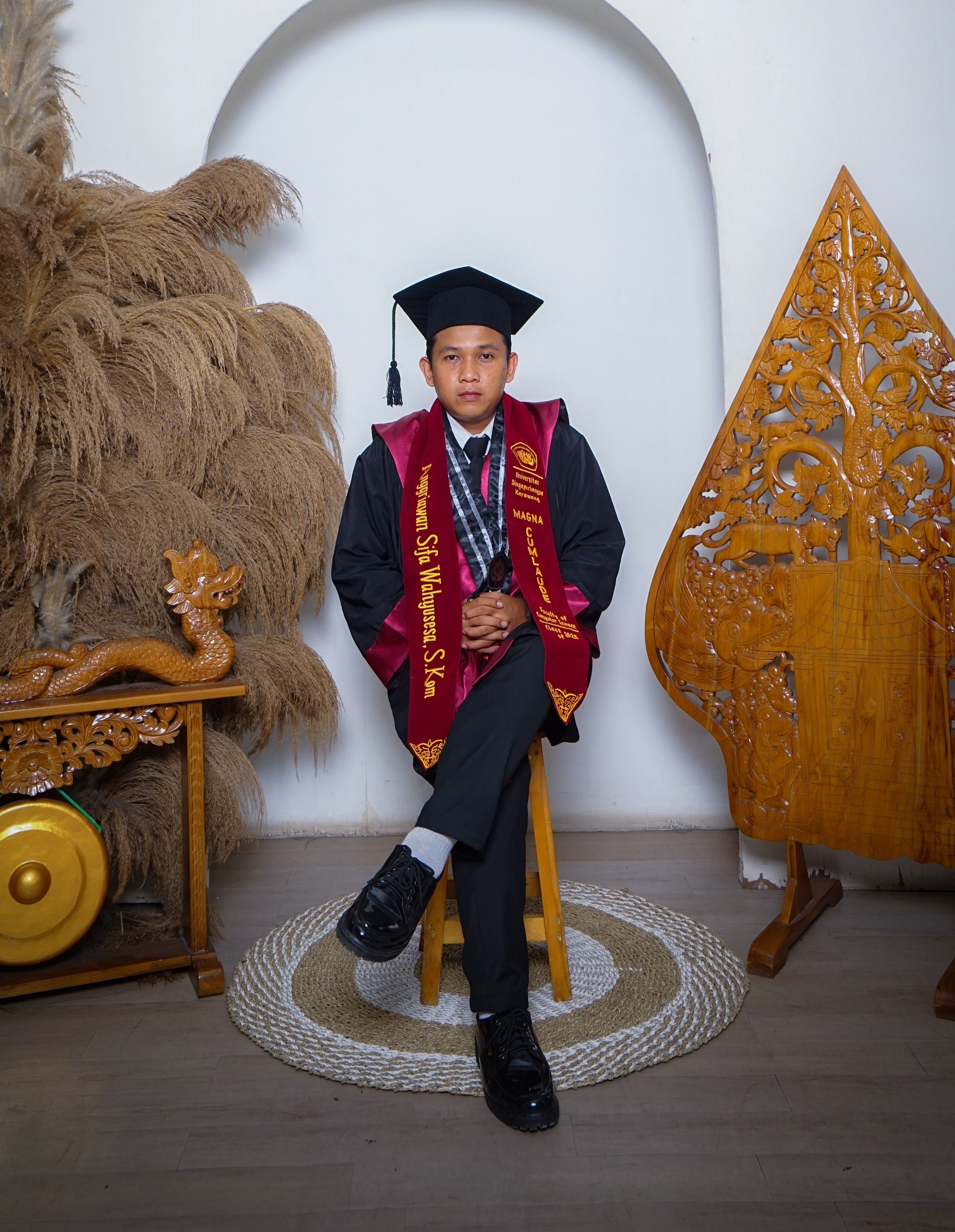 A young graduate in academic gown and cap sits surrounded by cultural decor indoors.