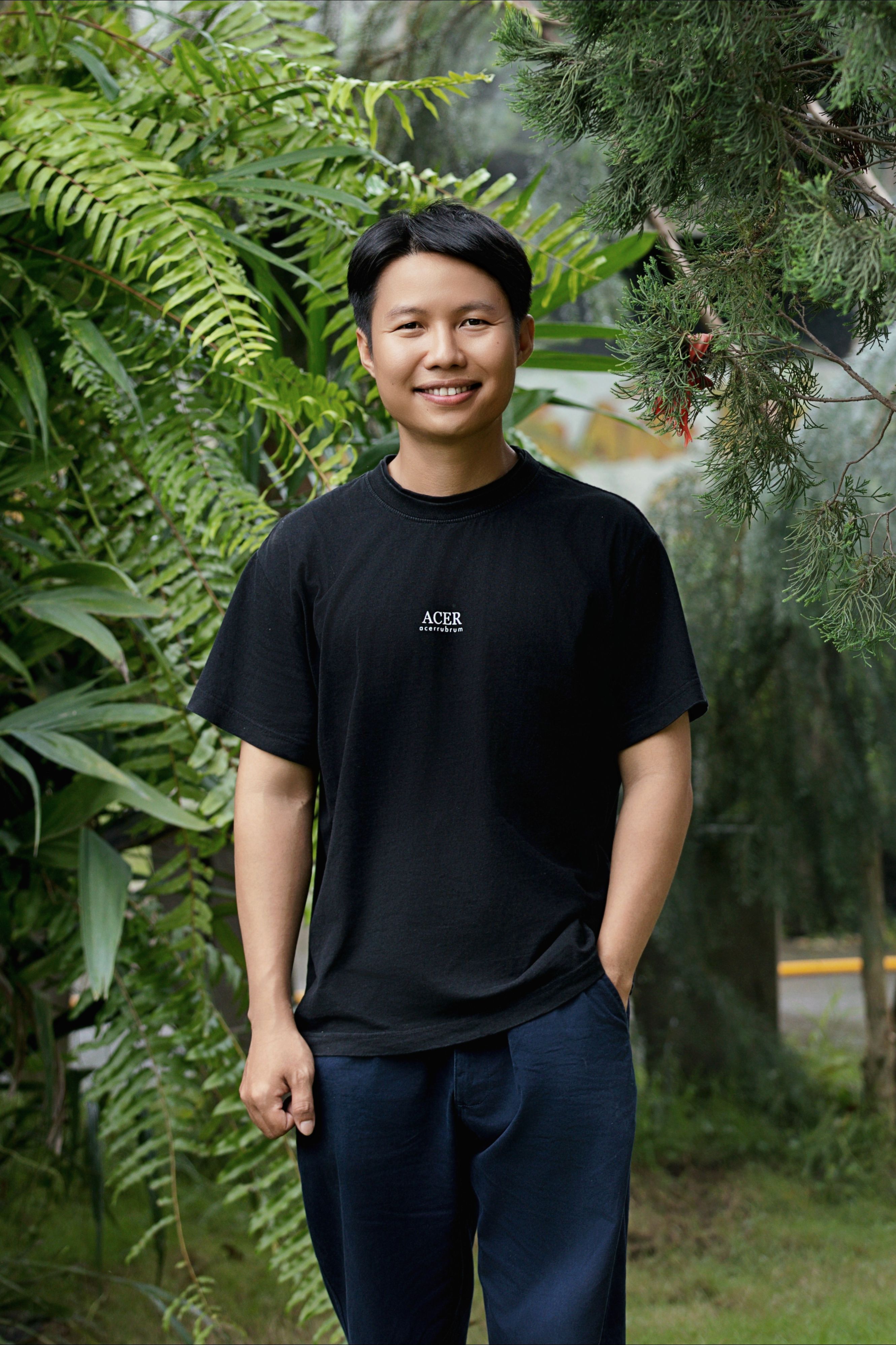 Young man in casual attire smiling amidst lush green plants outdoors.