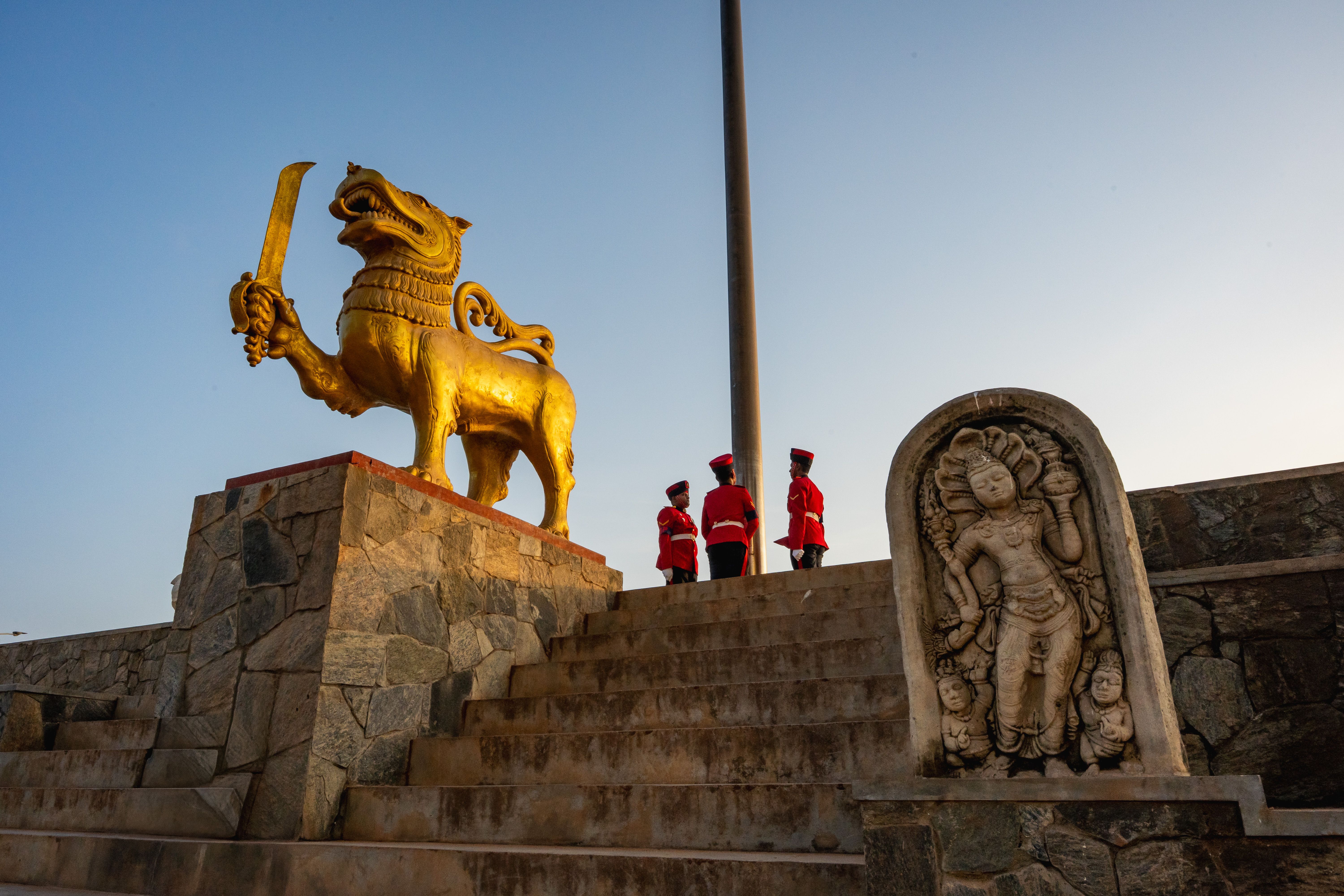 Three uniformed guards stand near the golden lion statue in Colombo at sunset.