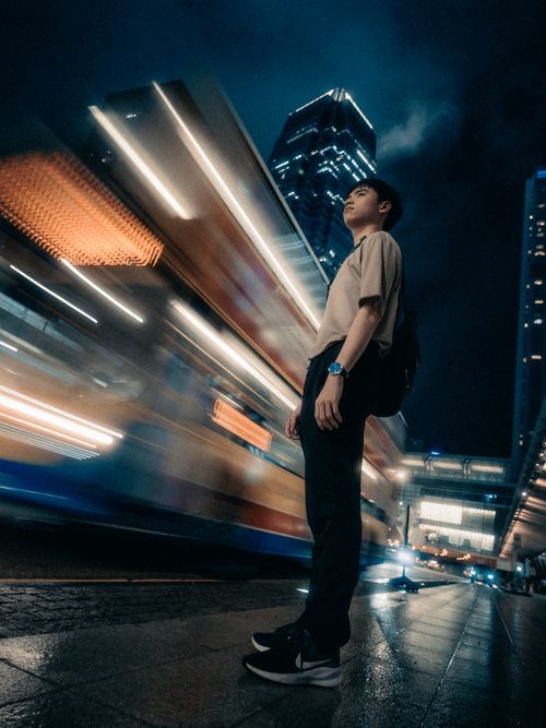 Free A young adult stands on a city street at night, with motion blurred lights of a passing bus. Stock Photo