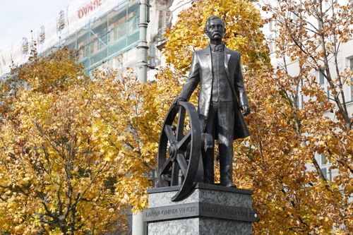 Free Bronze statue with an industrial wheel stands amidst vibrant autumn trees in an urban setting. Stock Photo