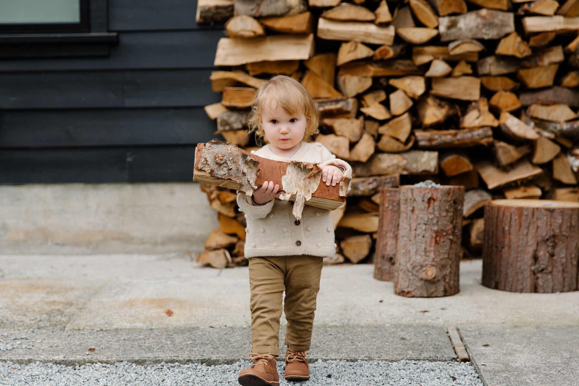 Charming scene of a child carrying firewood outside by a stack of logs, exuding cuteness.