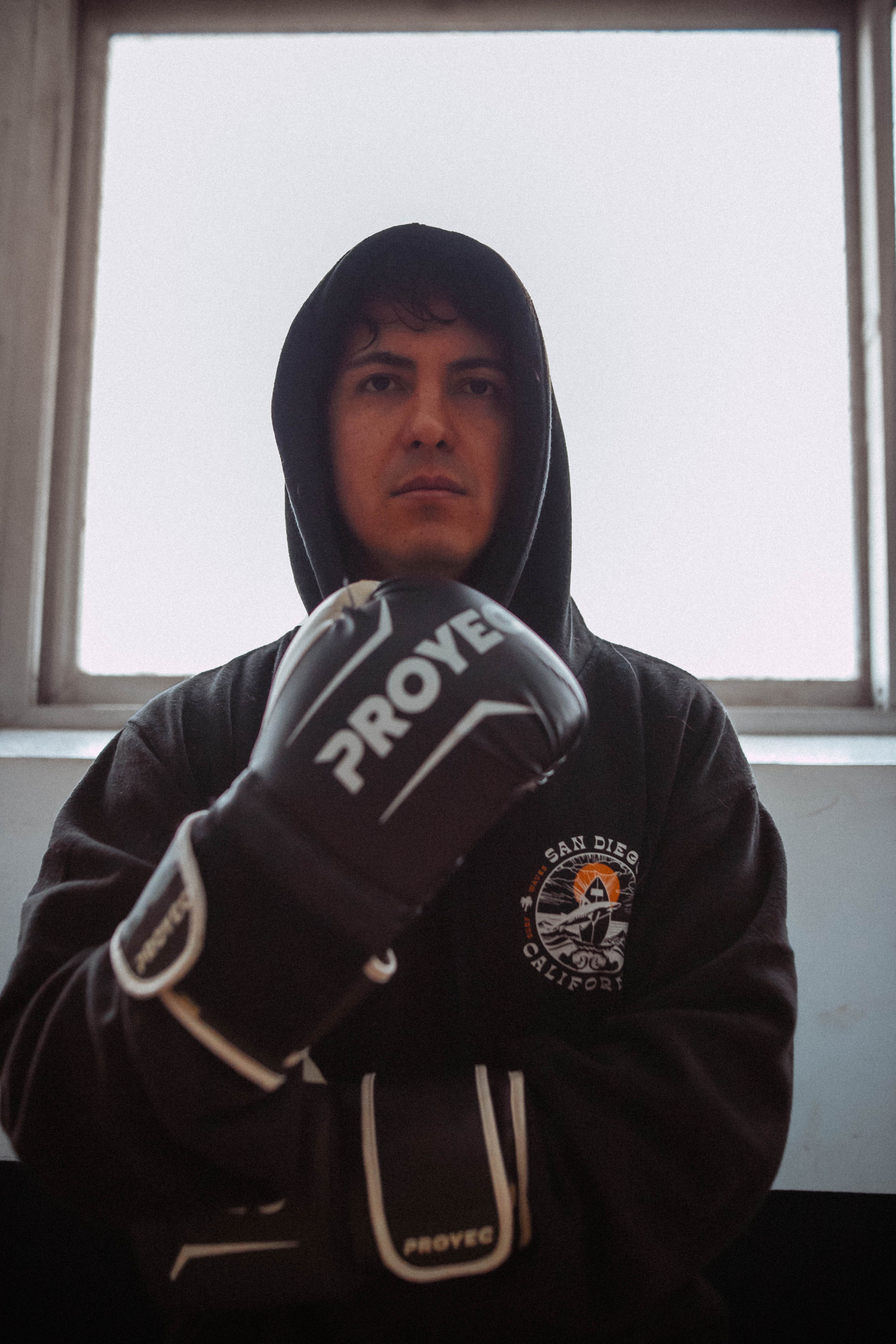 A male boxer wearing gloves and a hoodie poses indoors in front of a window.
