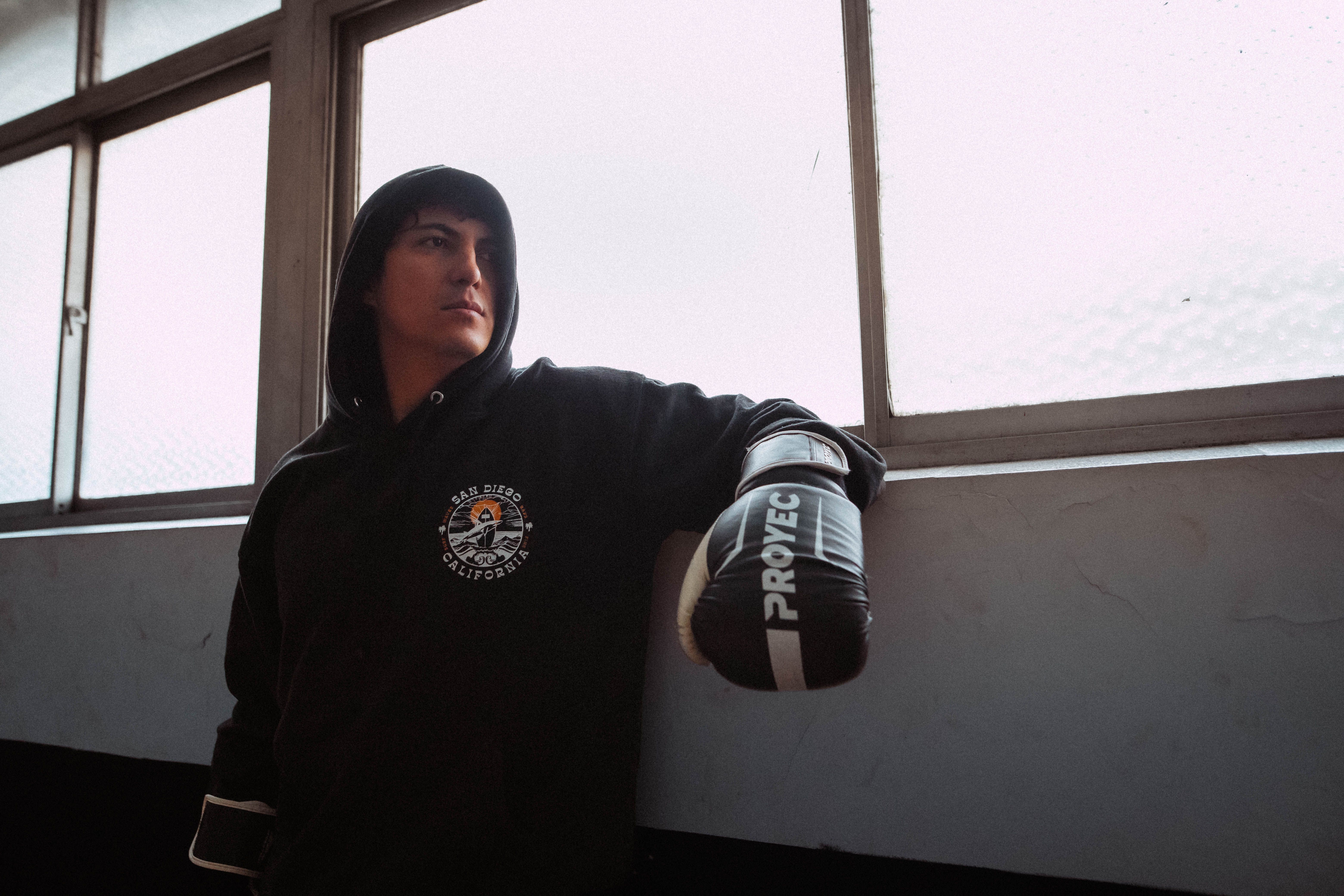 A boxer wearing a black hoodie and gloves leans against a wall indoors, exuding determination.