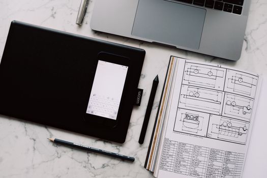 A modern workspace featuring a drawing tablet, smartphone, laptop, and design documents on a marble desk.