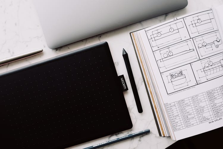 Graphic Tablet And Open Book With Drawing Located On Marble Table