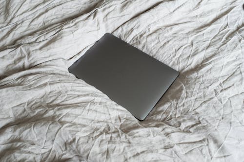 From above of closed dark gray portable computer located on large soft and crumpled cotton bed sheet in apartment in light room