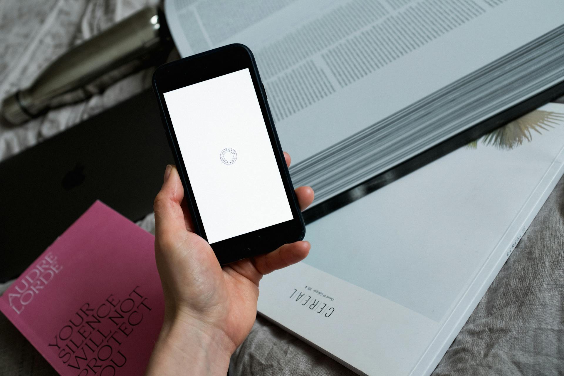 A close-up of a hand holding a smartphone displaying a loading screen over books and a laptop.