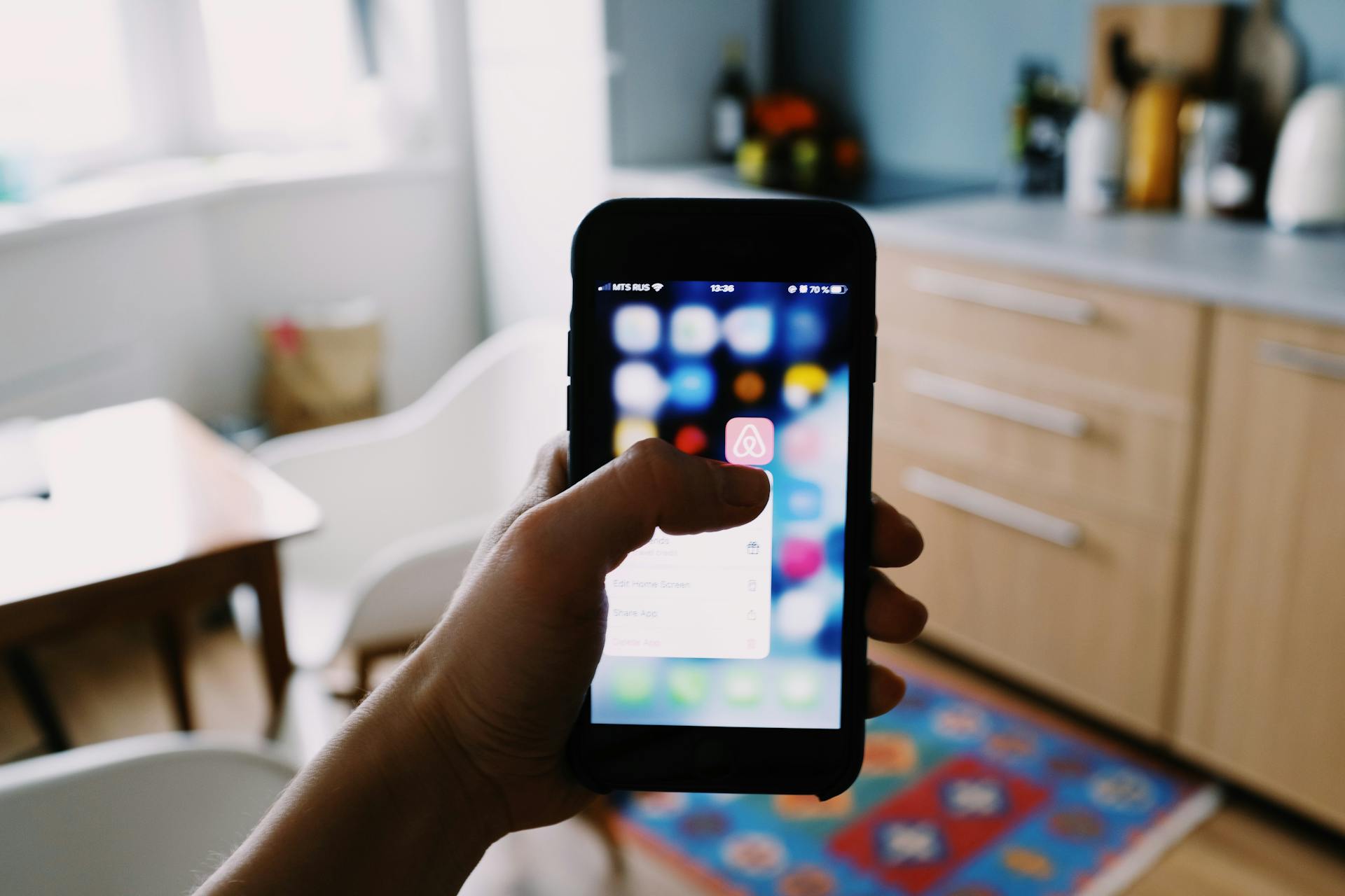 Crop anonymous person demonstrating app touching mobile phone screen while taking gadget with hand in kitchen in apartment in sunlight