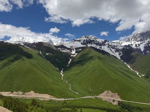 Free Panoramic landscape of lush green hills and snow-covered peaks under a bright blue sky. Stock Photo