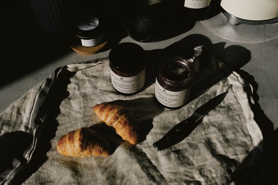 A cozy breakfast scene with fresh croissants, jars of jam, and a knife on a linen cloth illuminated by natural light.