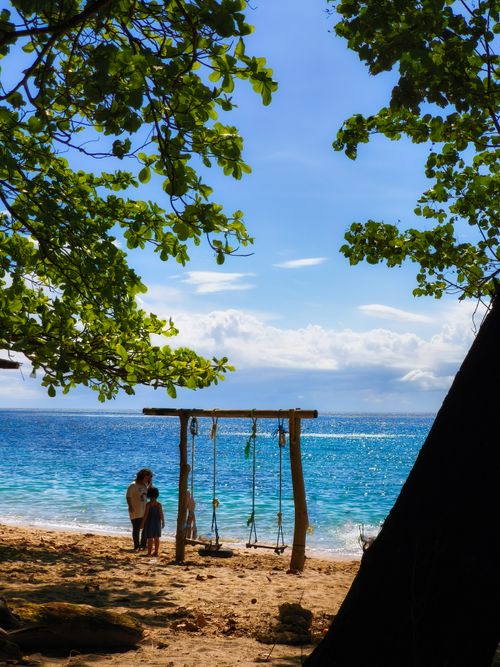 Free A serene tropical beach scene with swings, trees, and crystal-clear ocean waters under a bright sky. Stock Photo