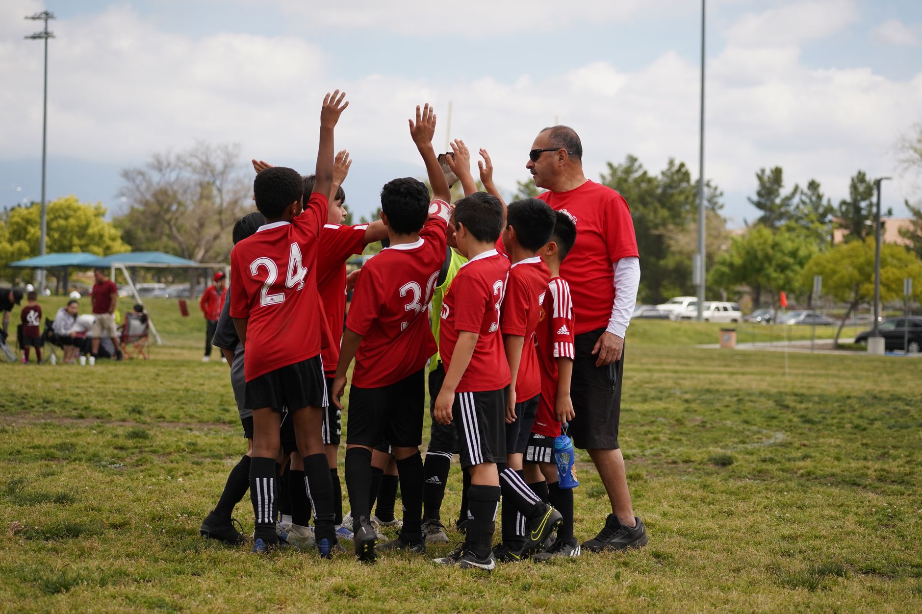 A youth soccer team in red jerseys gathers in a huddle with their coach on an outdoor field.