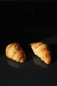 Two freshly baked croissants on a reflective dark surface with dramatic lighting.