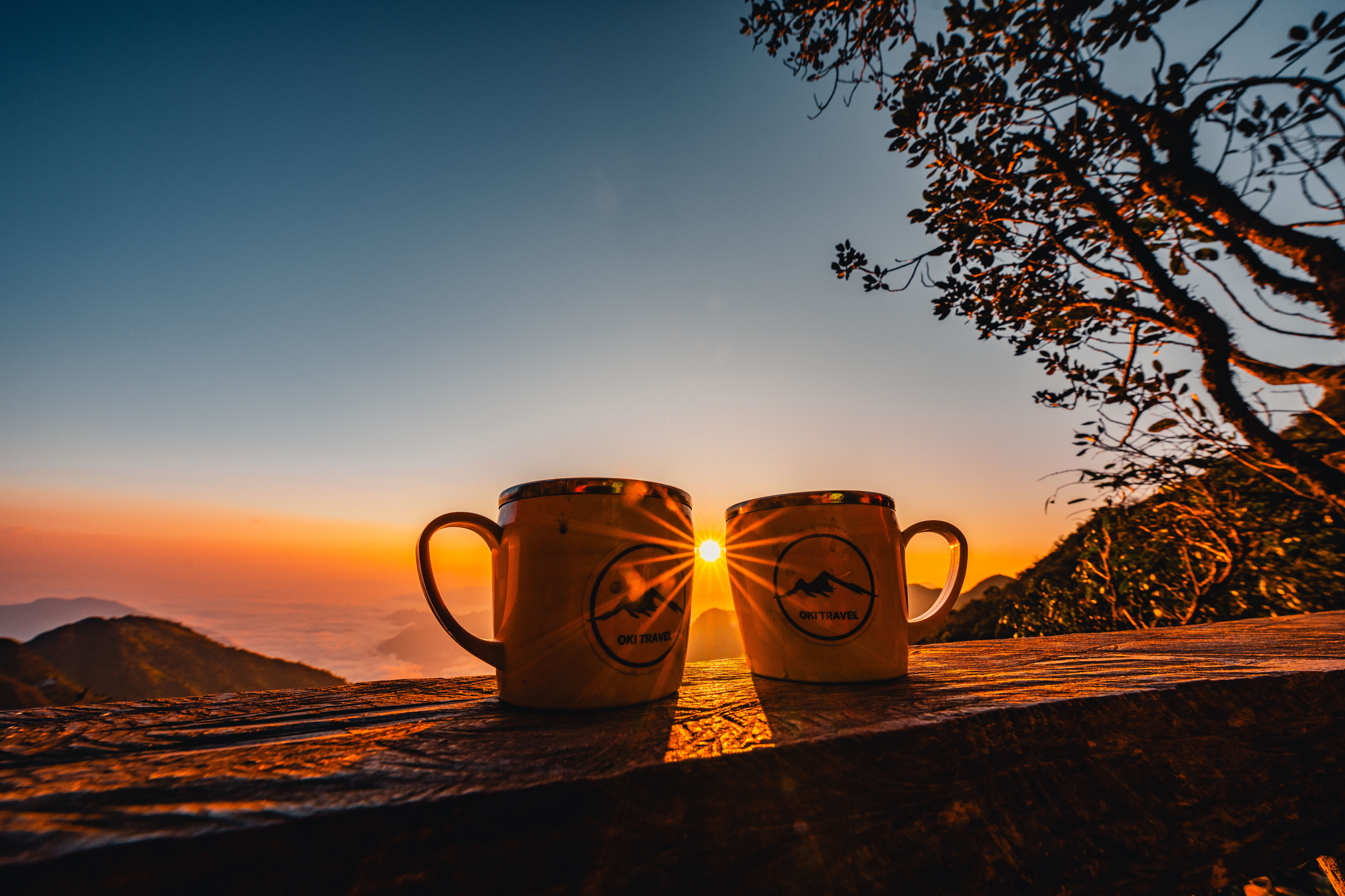 Golden sunrise over mountains with two camping mugs on a wooden bench, serene outdoor scene.