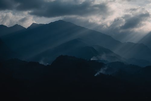 Free Majestic mountain landscape with dramatic clouds and sun rays piercing through, creating a moody atmosphere. Stock Photo