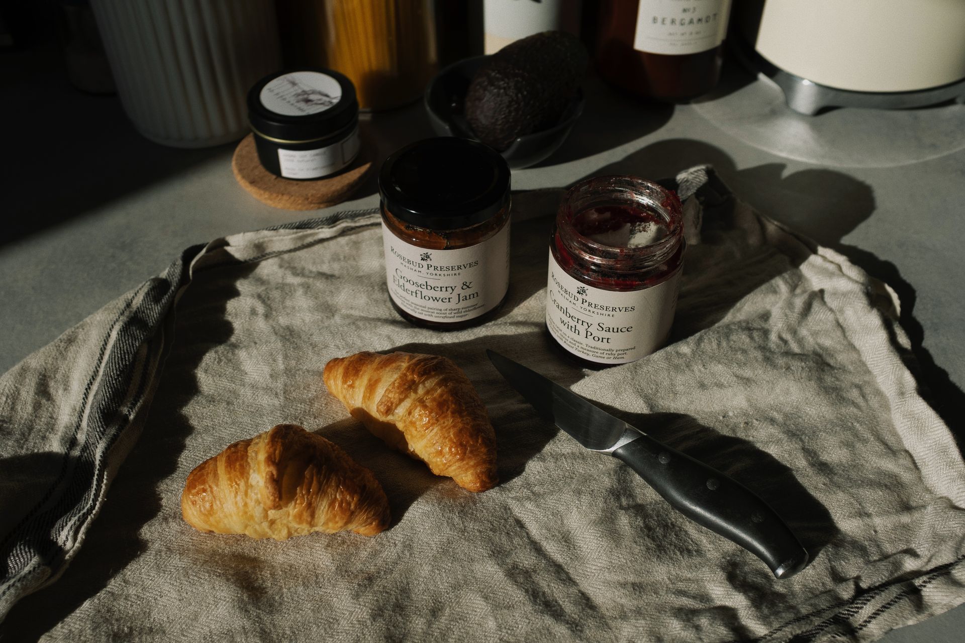 Still life of delicious brown croissants with opened jam and sauce pots on gray kitchen counter placed on with fabric napkin near knife in rustic style