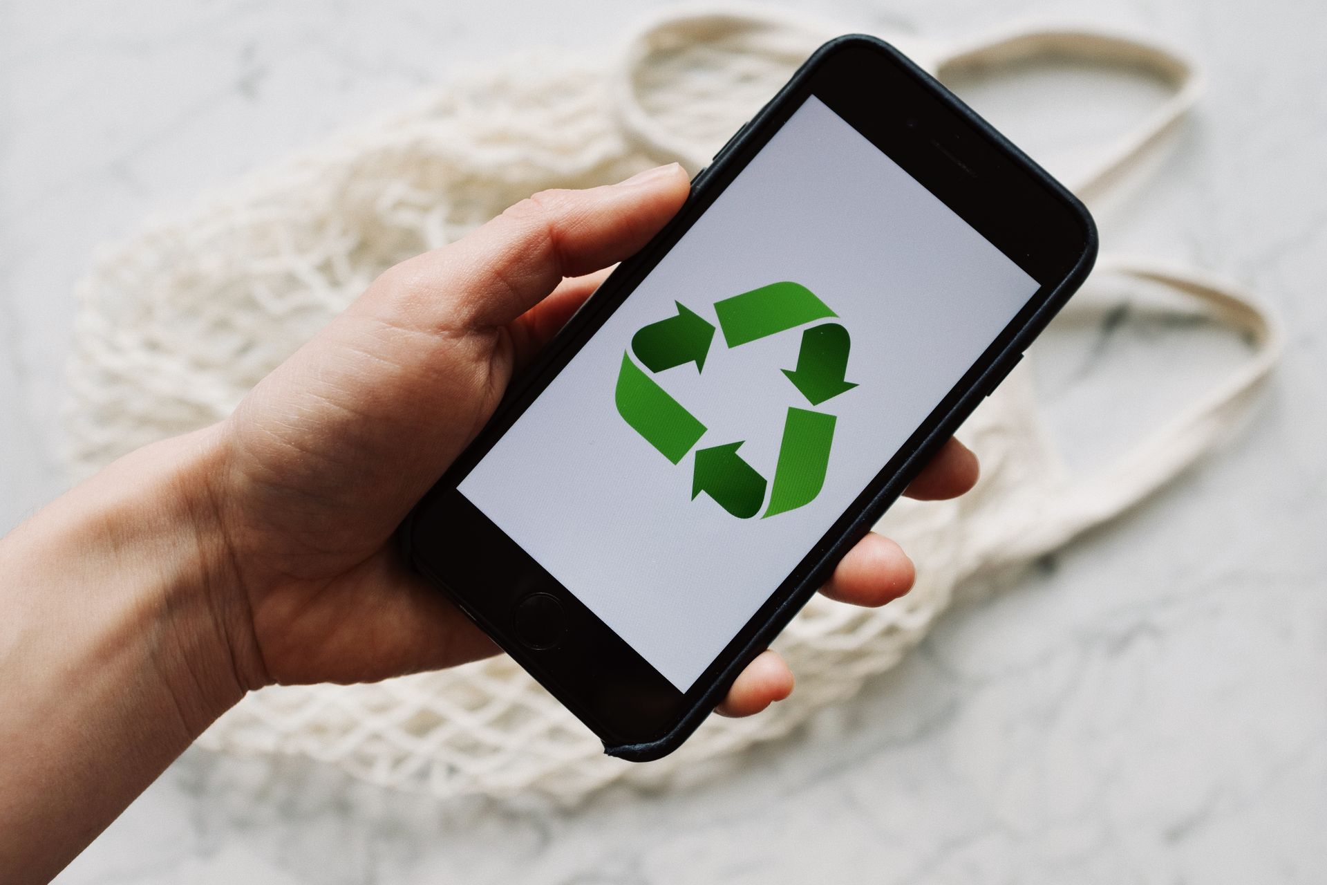 A smartphone with a recycling symbol held above a white net bag, emphasizing eco-friendly concepts.
