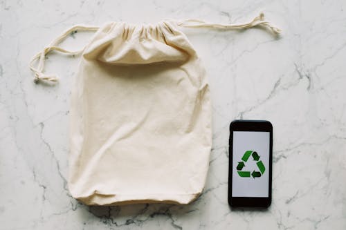 Free From above view of eco friendly cotton sack with drawstrings and smartphone with green recycling symbol on screen composed on white marble surface Stock Photo