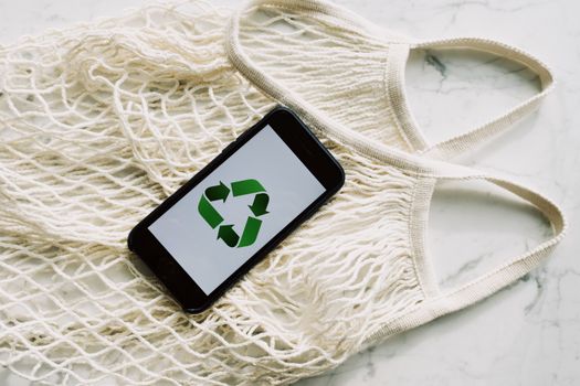 From above view of smartphone with green recycling icon on screen placed on empty cotton white mesh bag on marble table in light room