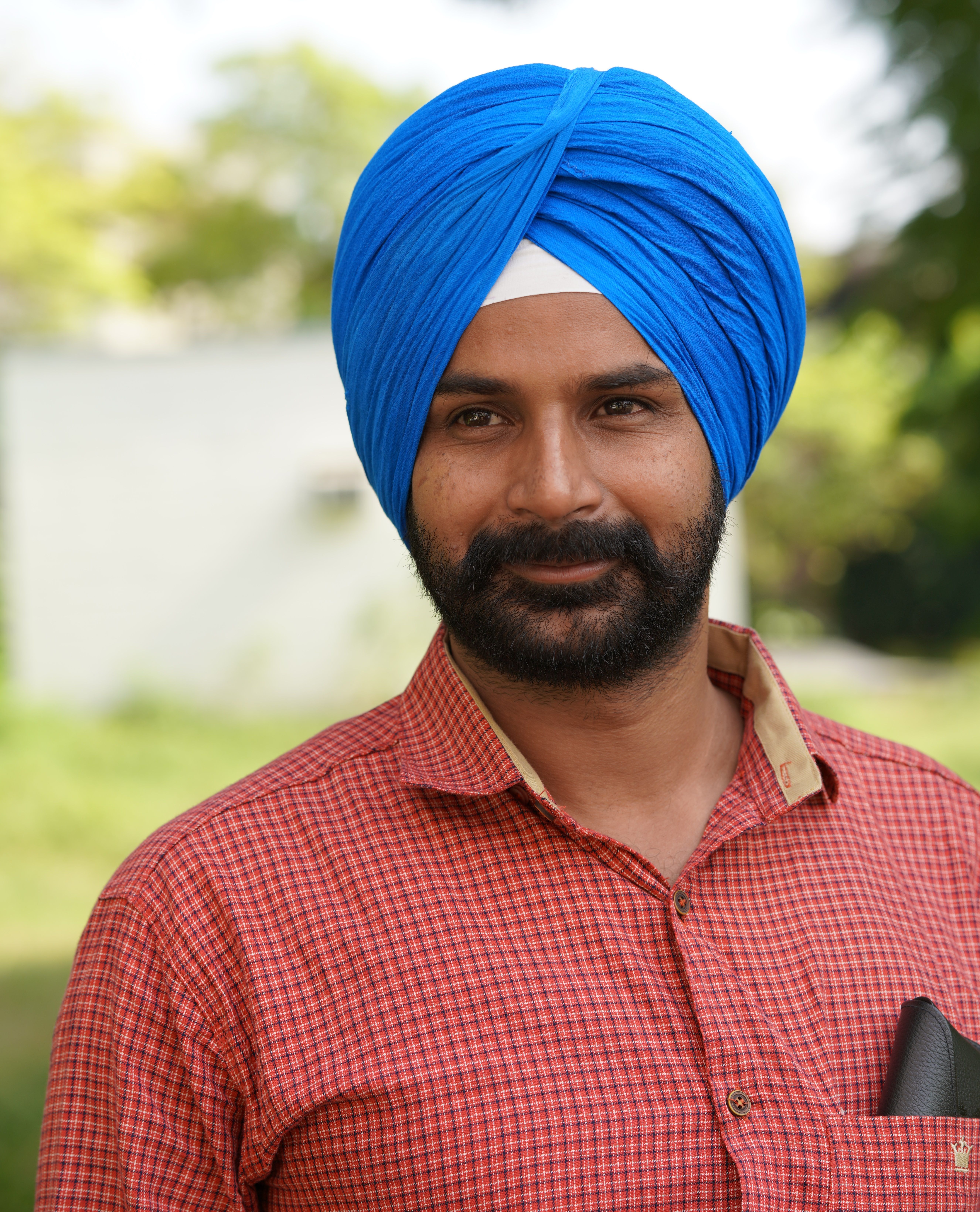 Close-up portrait of a man wearing a blue turban and red checkered shirt, standing outdoors.