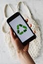 Top view of faceless person holding mobile phone with green recycle logo on white background above cotton eco mesh bag on marble table