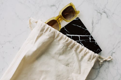 From above view of modern sunglasses and marble notepad placed in plain white textile bag on marble white table