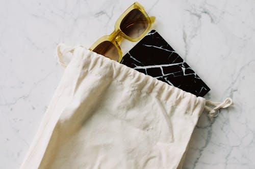 From above view of stylish sunglasses and book in black cover in white textile bag with drawstrings placed on marble surface in room