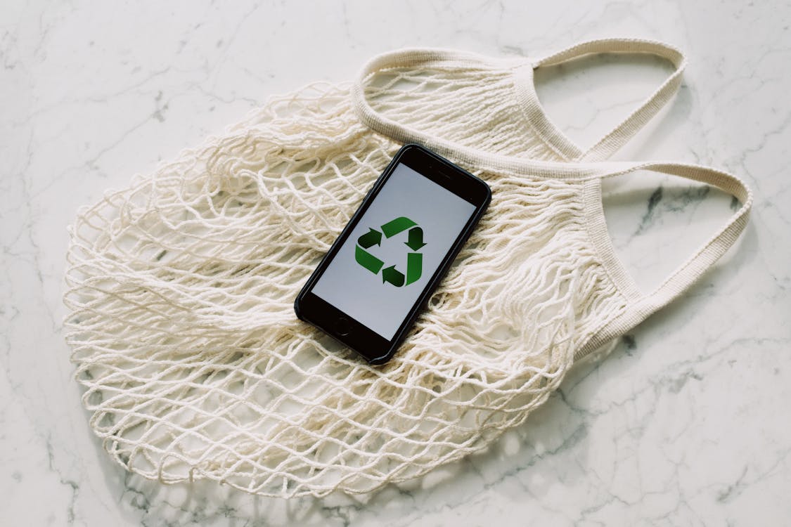 Overhead of smartphone with simple recycling sign on screen placed on white eco friendly mesh bag on marble table in room