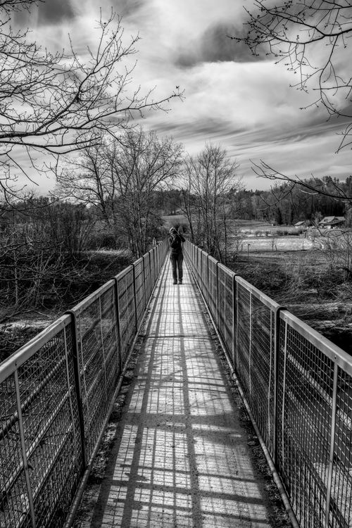 Free A lone person walking on a bridge amidst a serene Swiss landscape captured in black and white. Stock Photo