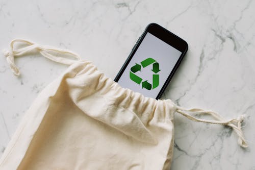Top view of plain white cotton sack and smartphone with green recycle emblem composed on marble surface