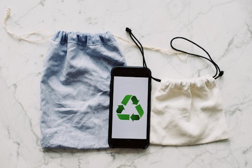 From above view of smartphone with recycling symbol on screen placed between white and blue textile bag on marble table in room