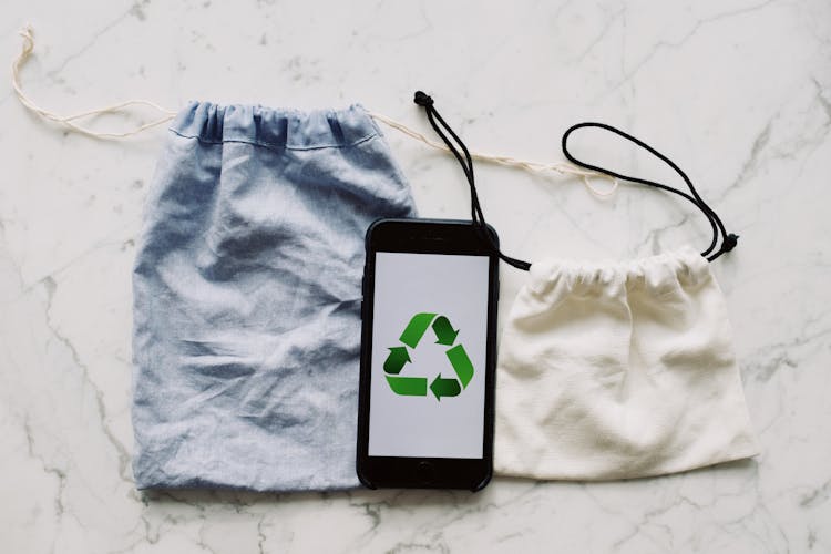 Smartphone With Recycling Symbol On Screen Placed Between White And Blue Textile Bag On Marble Table