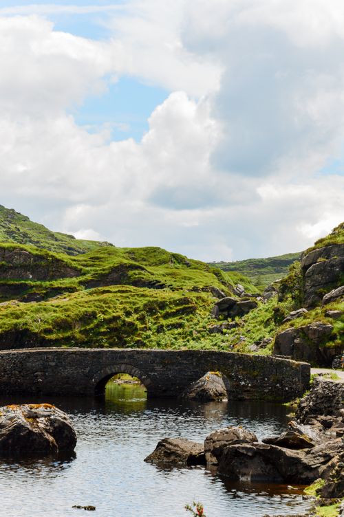 Free Picturesque stone bridge over a tranquil stream with lush greenery under a cloudy blue sky in Ireland. Stock Photo